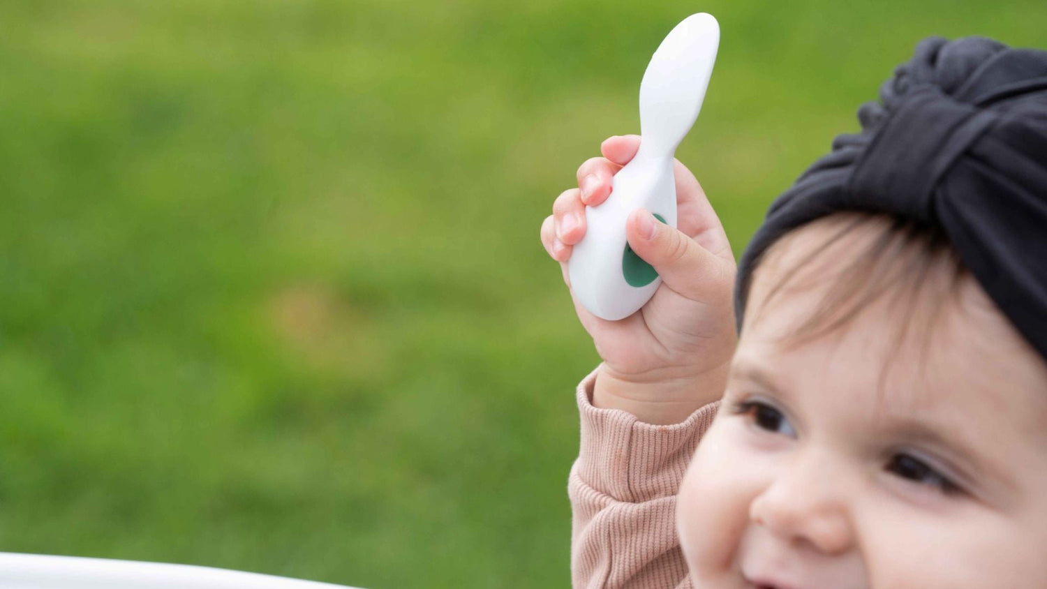 Smiling baby girl holding a doddl baby spoon triumphantly in her hand. The background is a garden lawn.