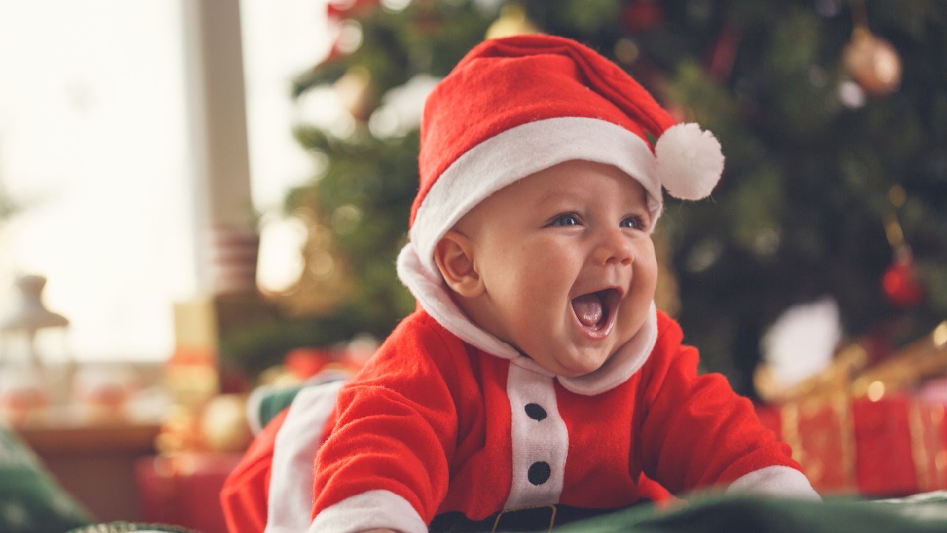 happy baby dressed in a santa outfit in front of a Christmas tree, surrounded by gifts