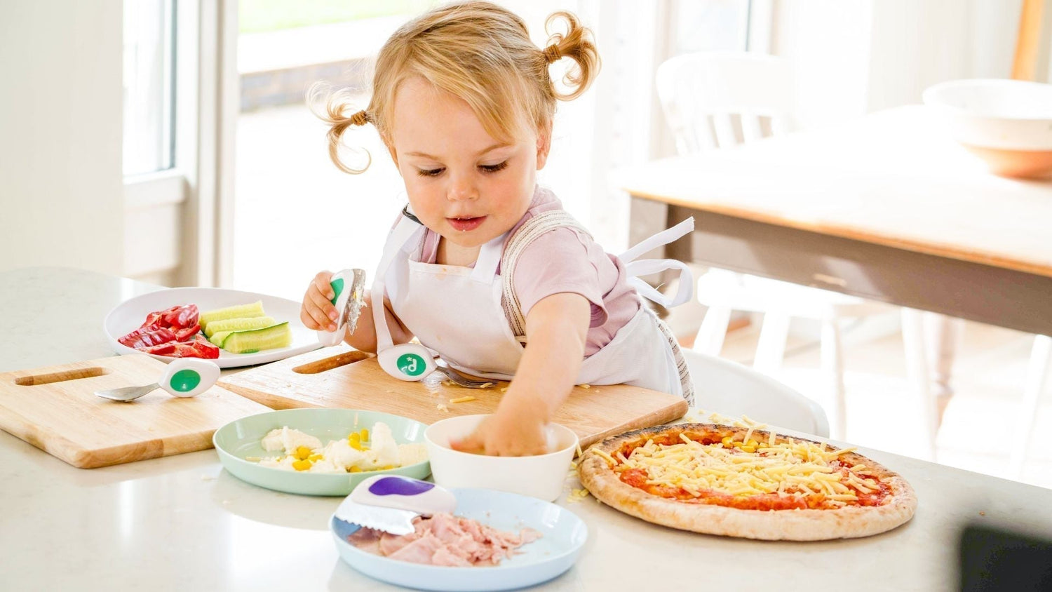 happy little girl uses her doddl cutlery to prepare a pizza, with lots of bowls and choices for pizza toppings on the clean white kitchen surface in front of her