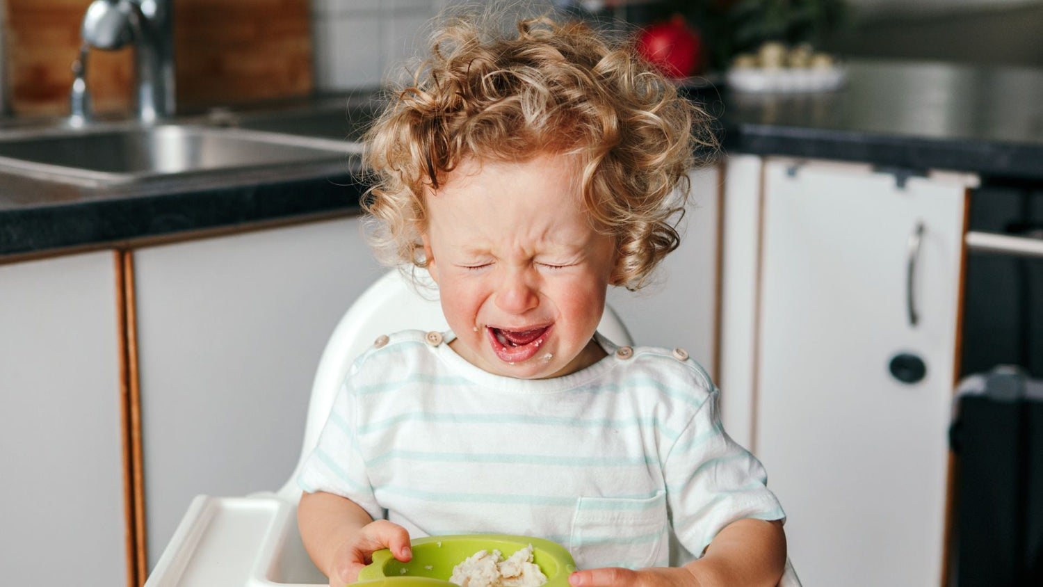 Toddler sitting in a highchair with a meal in front of them, looking angry and crying
