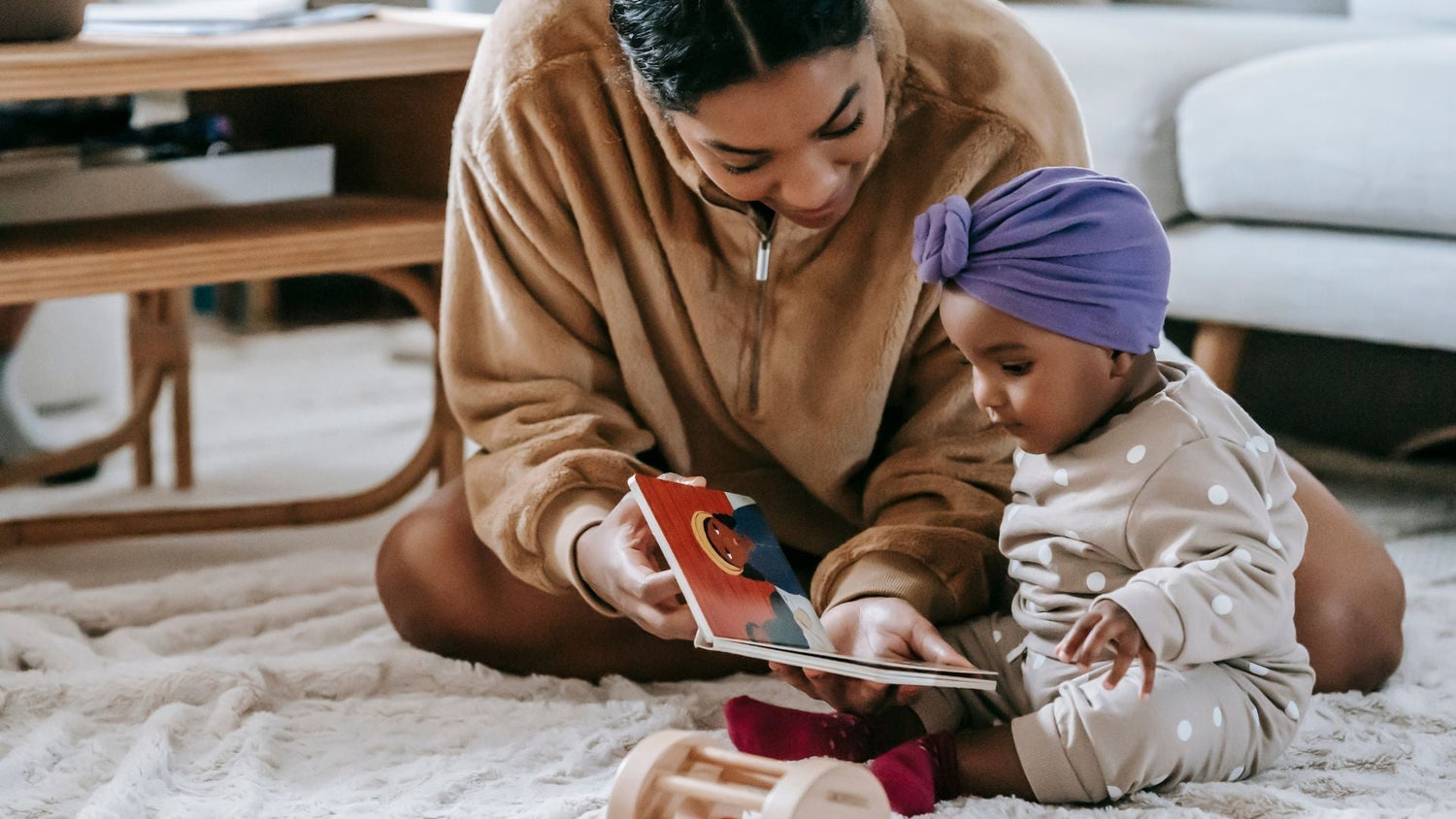 a mother sits on the floor reading a storybook to her baby. the mum is smiling and the baby is clearly transfixed by the book