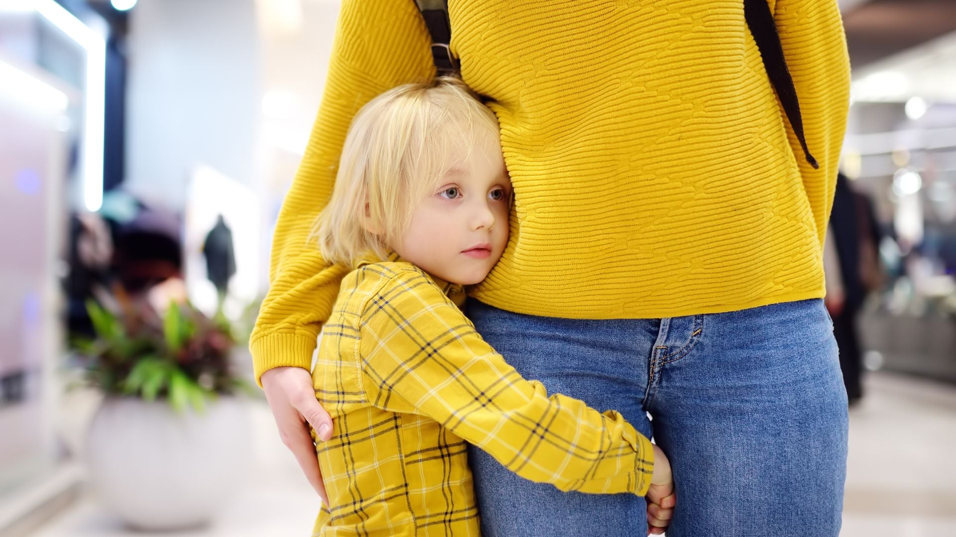 Small child in a yellow shirt looks sad and clings onto the leg of the female adult, who is wearing blue jeans and a yellow jumper