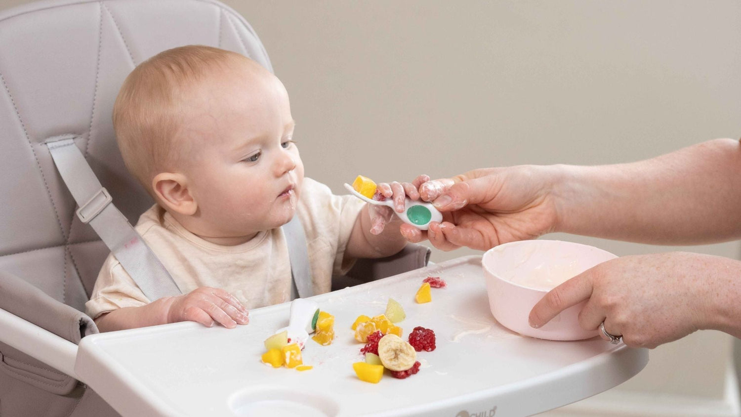 Baby boy sitting at a high chair, looking with interest at fingere foods and fruit offered on a doddl spoon