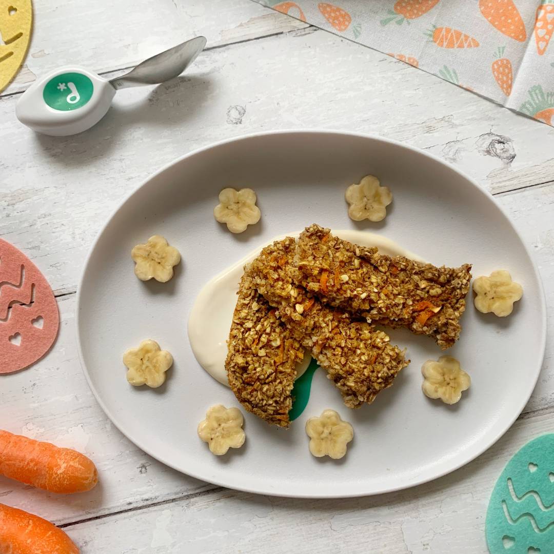 Carrot cake porridge fingers. 3 porridge fingers on a white doddl plate, with banana shapes and springtime colours in the background