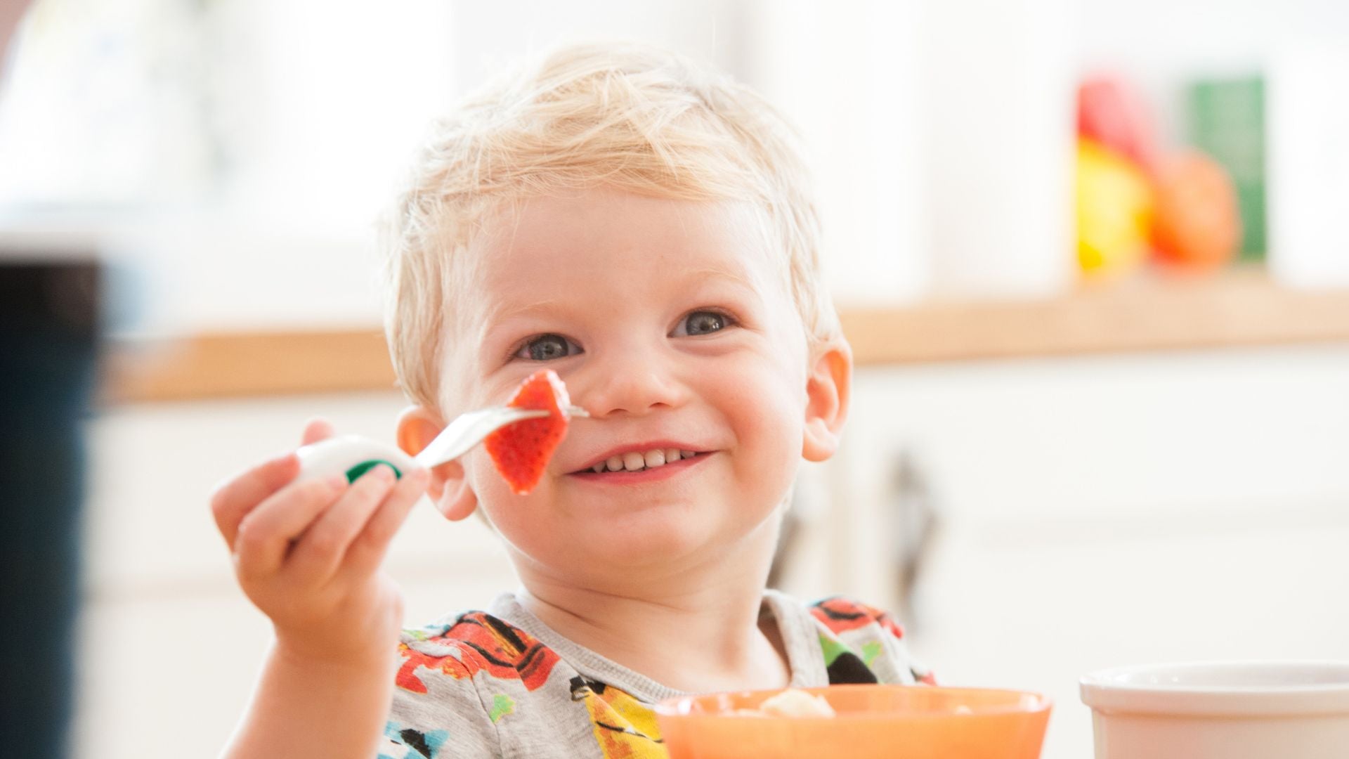 Happy toddler sitting at a table holding a piece of strawberry on his doddl toddler frok