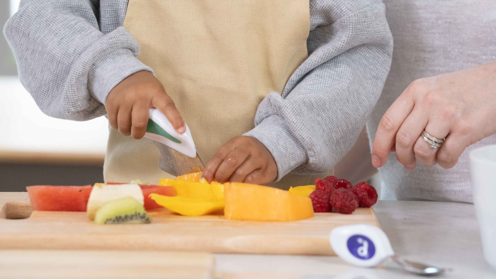 toddler hands holding a doddl kid-safe knife and cutting a selection of colourfulfruit