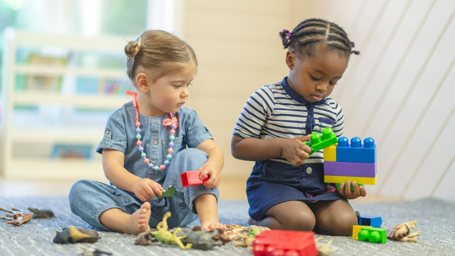 two toddlers sit on  carpet playing side by side with blocks