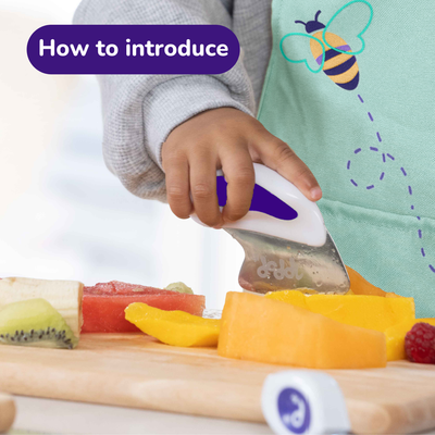 Child's hand using a doddl knife to cut fruit on a wooden board with a bee illustration in the corner.